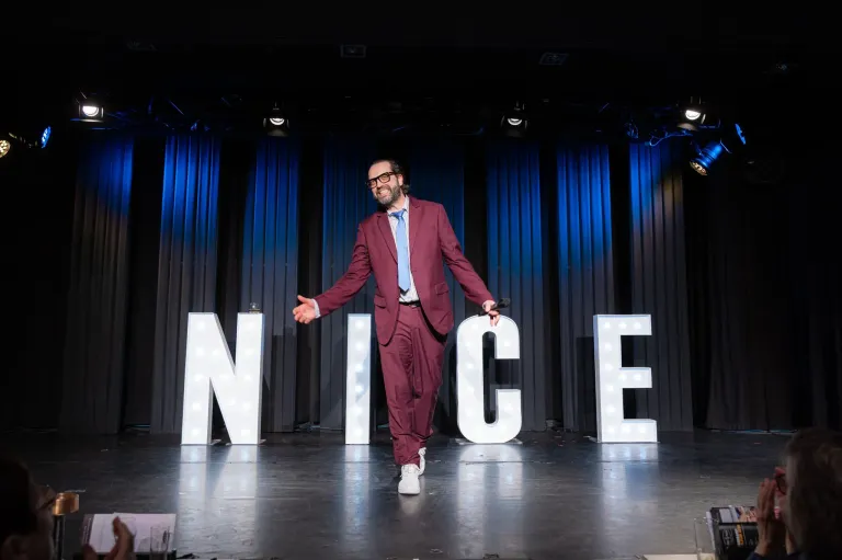 Ein Mann in einem roten Anzug steht lächelnd auf der Bühne, ein Mikrofon in der Hand, hinter großen weißen Buchstaben, die NICE schreiben.