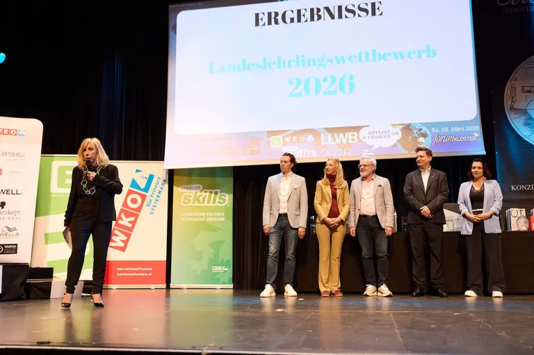 Fünf Personen, wahrscheinlich ein Team, stehen auf der Bühne. Dahinter befindet sich eine große Leinwand mit Text auf Deutsch. Die Frau links hält ein Mikrofon. Die Bühne ist dunkel und der Raum gut beleuchtet.