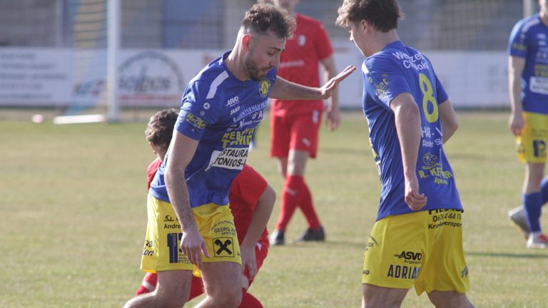 Ein Fußballspieler in Blau mit der Nummer 11 deutet auf einen anderen Spieler in Blau mit der Nummer 8 hin. Sie befinden sich auf einem Rasenfeld, und es gibt einen verschwommenen Spieler in Rot hinter ihnen.