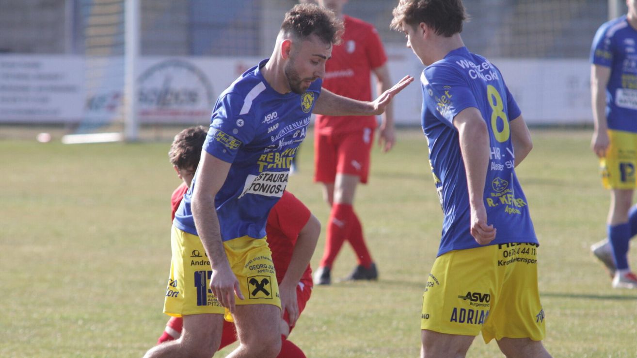Ein Fußballspieler in Blau mit der Nummer 11 deutet auf einen anderen Spieler in Blau mit der Nummer 8 hin. Sie befinden sich auf einem Rasenfeld, und es gibt einen verschwommenen Spieler in Rot hinter ihnen.