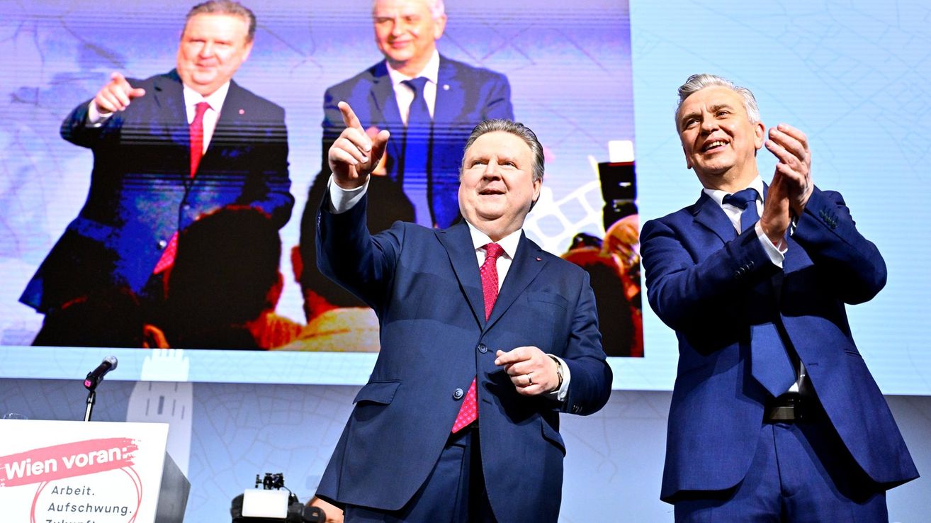 Drei Männer in Anzügen stehen auf einer Bühne mit einer großen Leinwand im Hintergrund. Der Mann in der Mitte zeigt auf etwas.