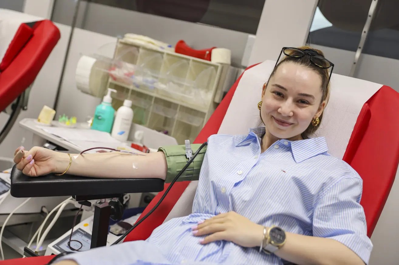 Eine Frau sitzt auf einem roten Stuhl und trägt ein blaues Hemd und eine grüne Manschette. Sie lächelt, wahrscheinlich in einem Blutspendezentrum. Hinter ihr steht ein weißer Regal mit verschiedenen Gegenständen.