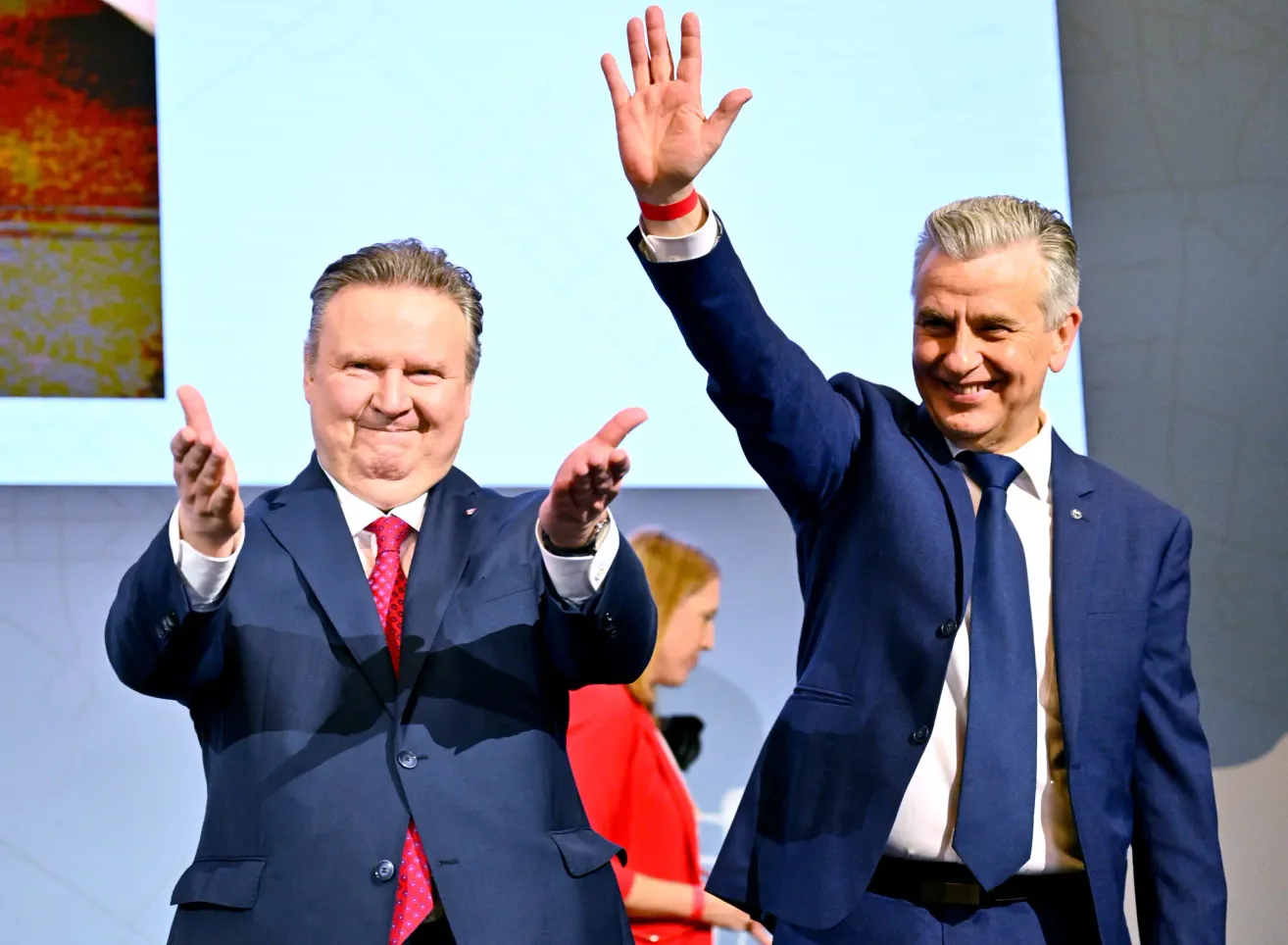 Zwei Männer in Anzügen winken in die Kamera. Ein Mann hat eine rote Krawatte, der andere ein rotes Armband. Hinter ihnen schaut eine Frau in einem roten Shirt zu ihnen.