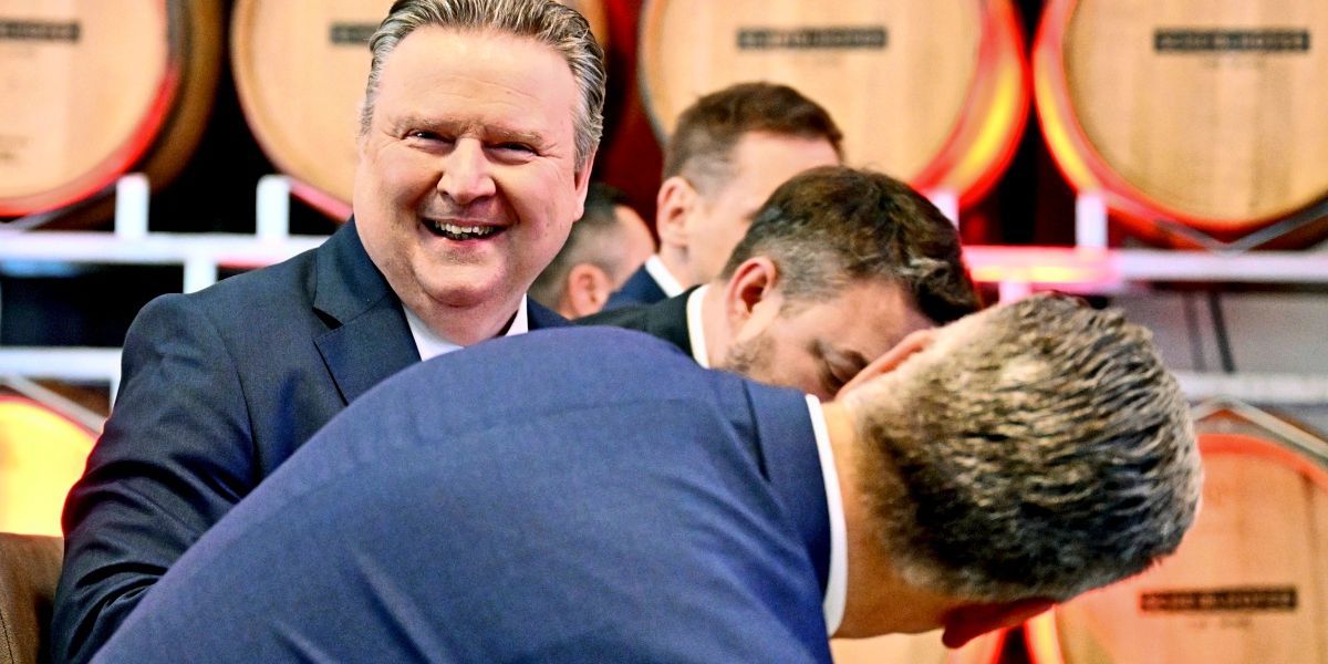 Ein Mann im Anzug lächelt fröhlich in einem Raum mit Holzfässern. Ein anderer Mann mit gesenktem Kopf ruht seinen Kopf auf seiner Schulter.