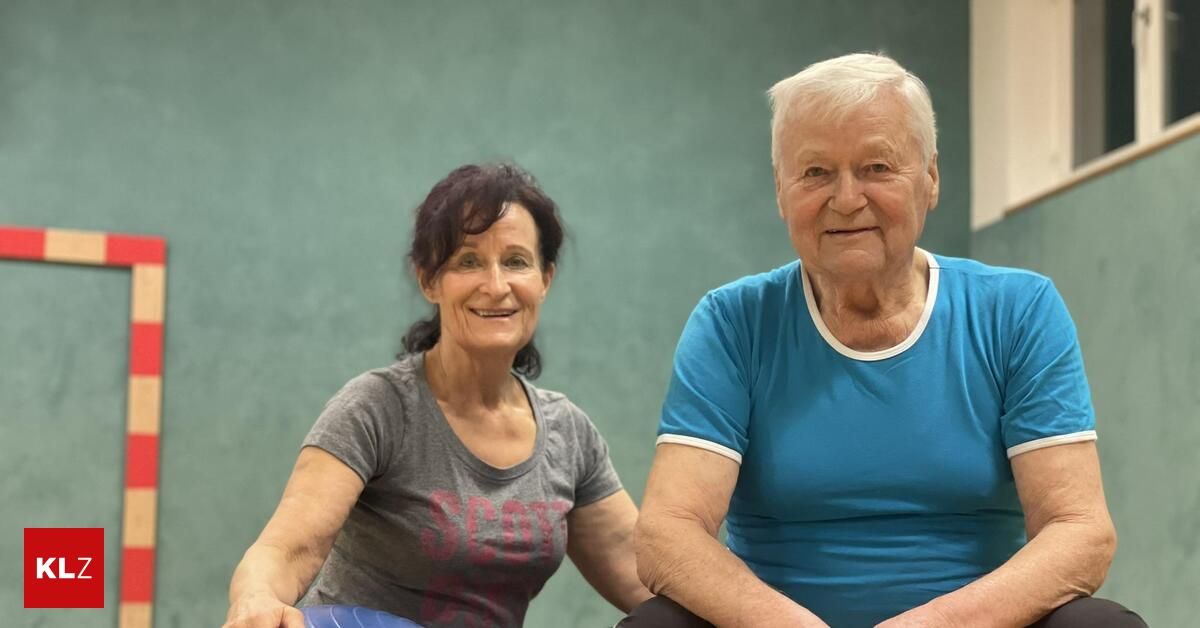 Zwei Menschen, eine Frau und ein älterer Mann, sitzen gemeinsam gegen eine grüne Wand, beide lächelnd. Die Frau trägt ein graues T-Shirt und der Mann ein blaues. Sie scheinen sich in einem Fitnessstudio oder einer Turnhalle zu befinden.