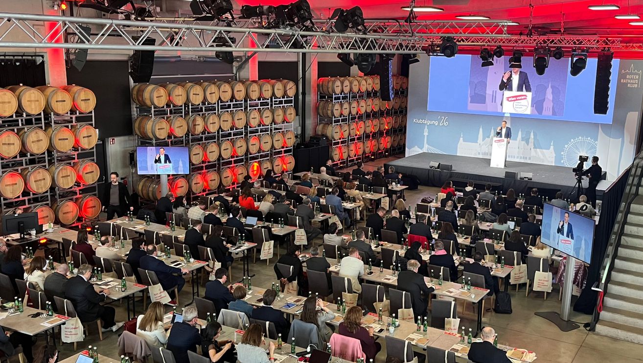 Ein Konferenzsaal mit vielen Menschen an Tischen, die einen großen Bildschirm auf der Bühne betrachten. Fässer sind hinter dem Publikum gestapelt, und ein Redner steht auf der Bühne.