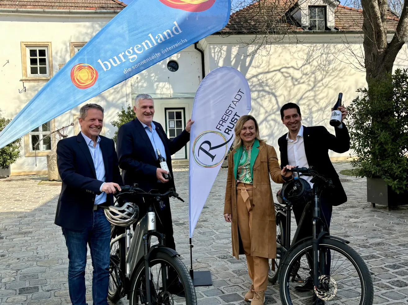 Vier Personen posieren mit E-Bikes vor einem Gebäude. Sie halten Flaschen in der Hand und scheinen zu lächeln. Ein Banner zeigt 'burgenland', ein anderes 'Freistadt Rust'.