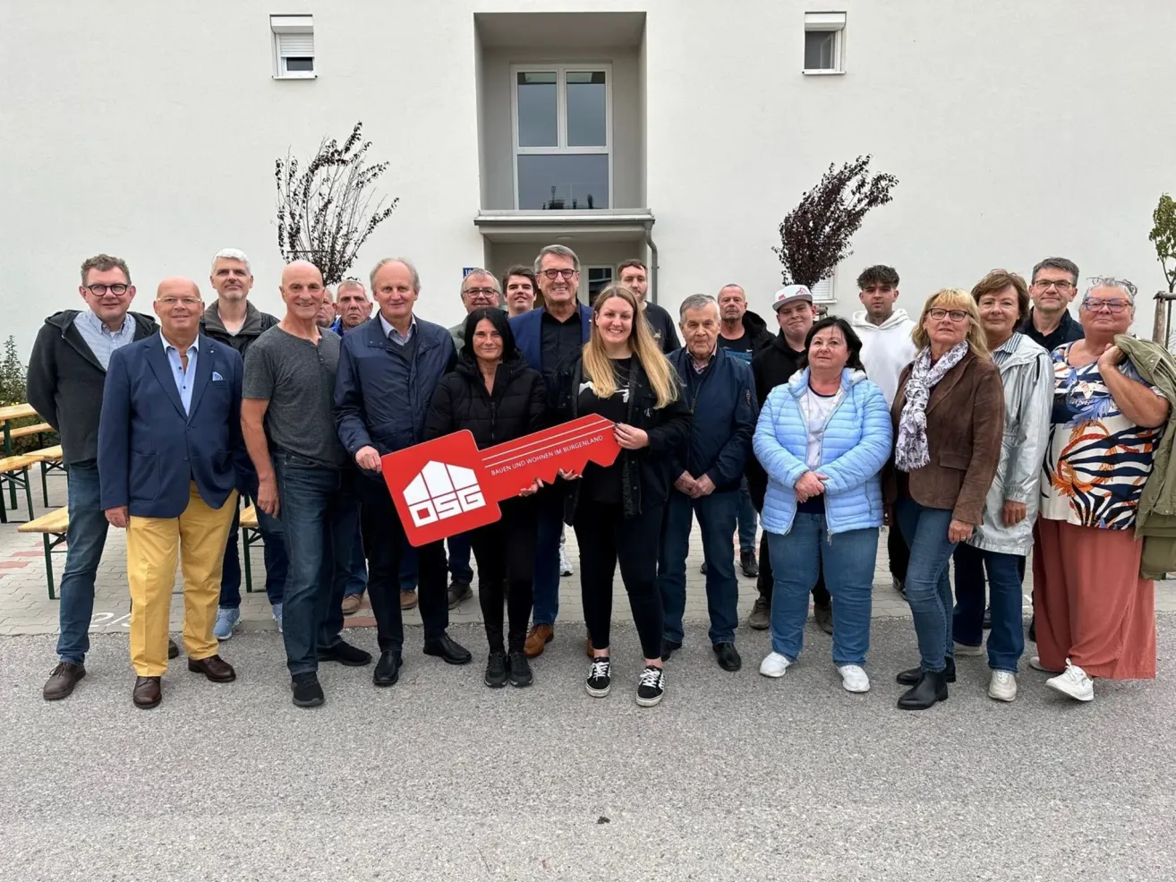 Eine Gruppe von Menschen posiert für ein Foto vor einem Gebäude und hält einen roten Schlüssel. Die Frau in der Mitte hält den Schlüssel mit beiden Händen.