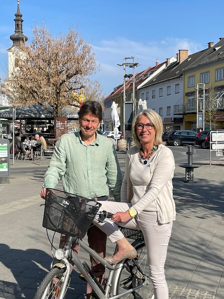 Ein Mann und eine Frau stehen neben einem Fahrrad mit Korb und lächeln in einer städtischen Umgebung. Hinter ihnen sind ein Baum, ein Gebäude mit Turm und geparkte Autos zu sehen.
