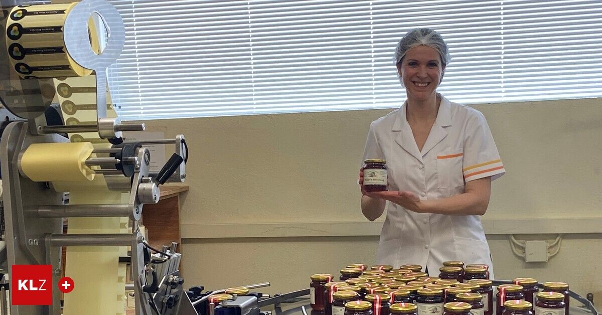 Eine Frau in einem weißen Kittel lächelt und hält ein Glas Marmelade in einer Küche mit Fenster und Maschine.