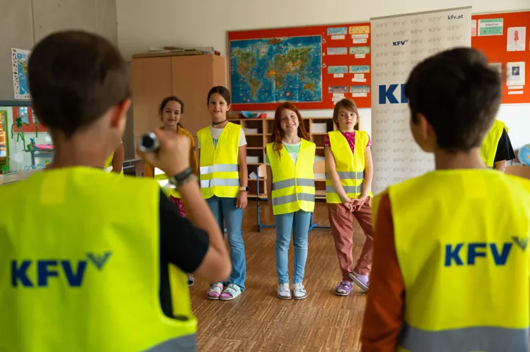 Fünf junge Mädchen in neonfarbenen Sicherheitswesten stehen in einem Klassenzimmer mit einer Karte im Hintergrund. Ein Junge macht ein Foto von ihnen.