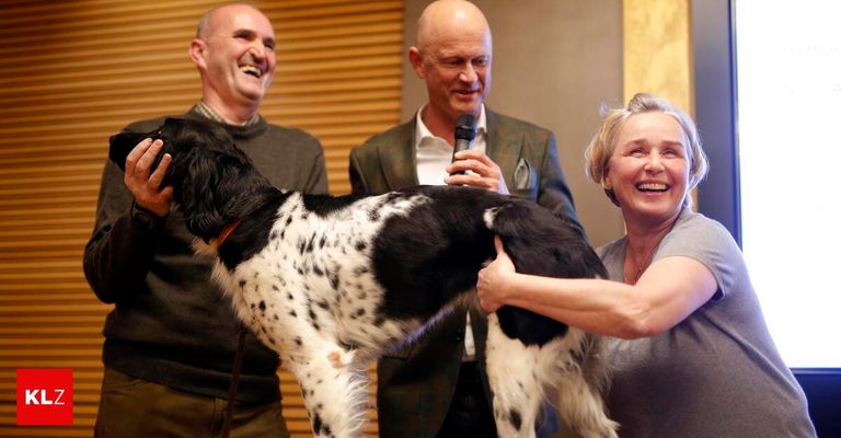 Drei Personen stehen mit einem Hund. Ein Mann hält ein Mikrofon und die Frau streichelt den Hund.