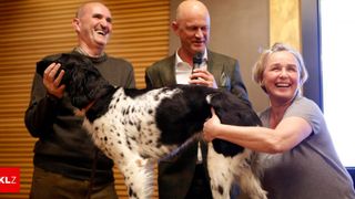 Drei Personen stehen mit einem Hund. Ein Mann hält ein Mikrofon und die Frau streichelt den Hund.