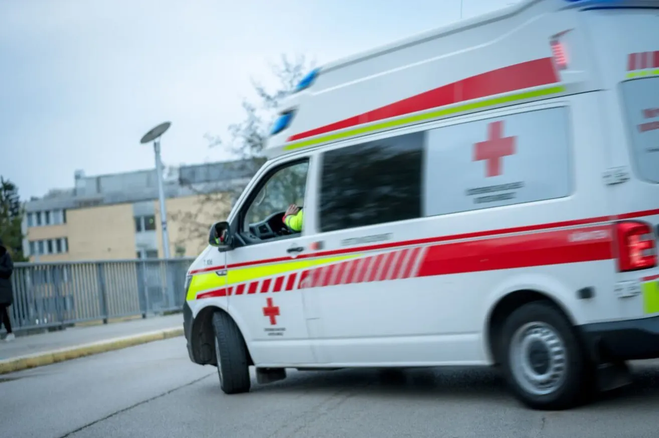 Ein Krankenwagen mit roten und gelben Streifen fährt eine Straße entlang. Der Fahrer trägt eine neongrüne Weste.