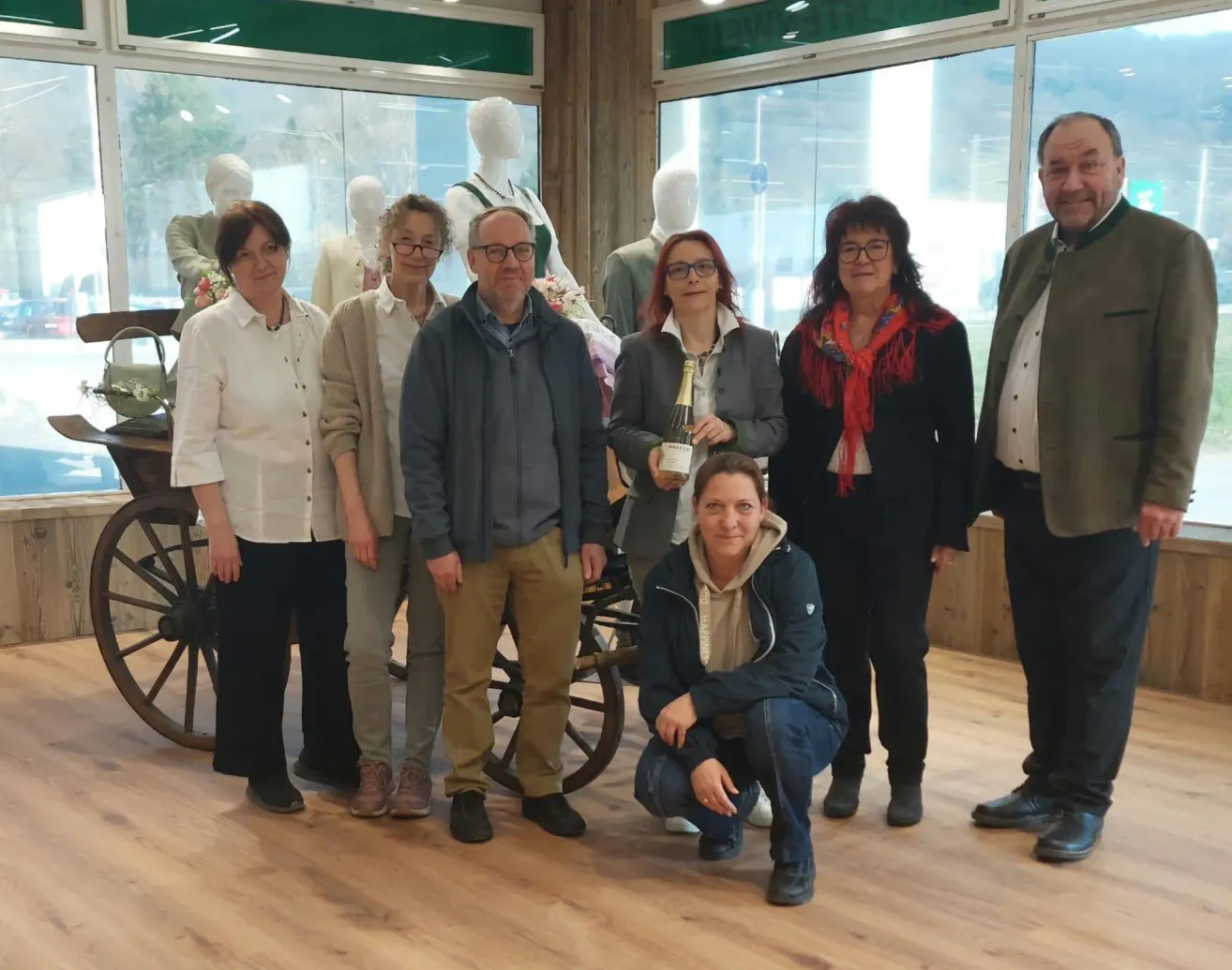 Eine Gruppe Erwachsener steht vor einer Glaswand mit Mannequins und hält eine Flasche Wein. Die Frau in der Mitte kniet und lächelt. Sie sind drinnen, mit Holzböden.