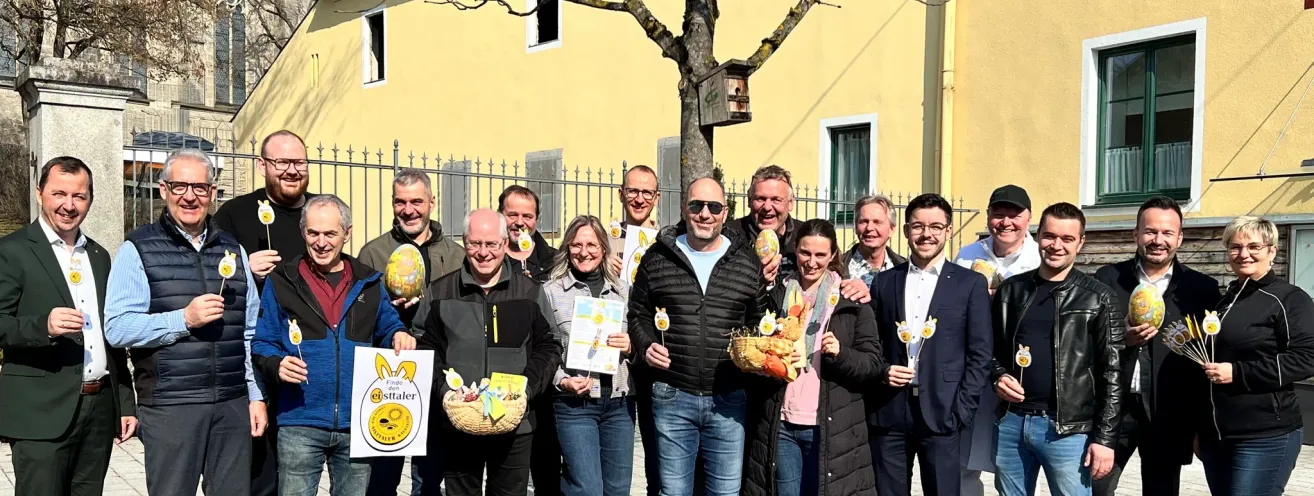 Eine Gruppe von Erwachsenen posiert für ein Foto vor einem Baum, sie halten Körbe mit Ostereiern und eine Frau hat ein Schild. Dahinter steht ein Gebäude mit einem Fenster und einem Zaun.