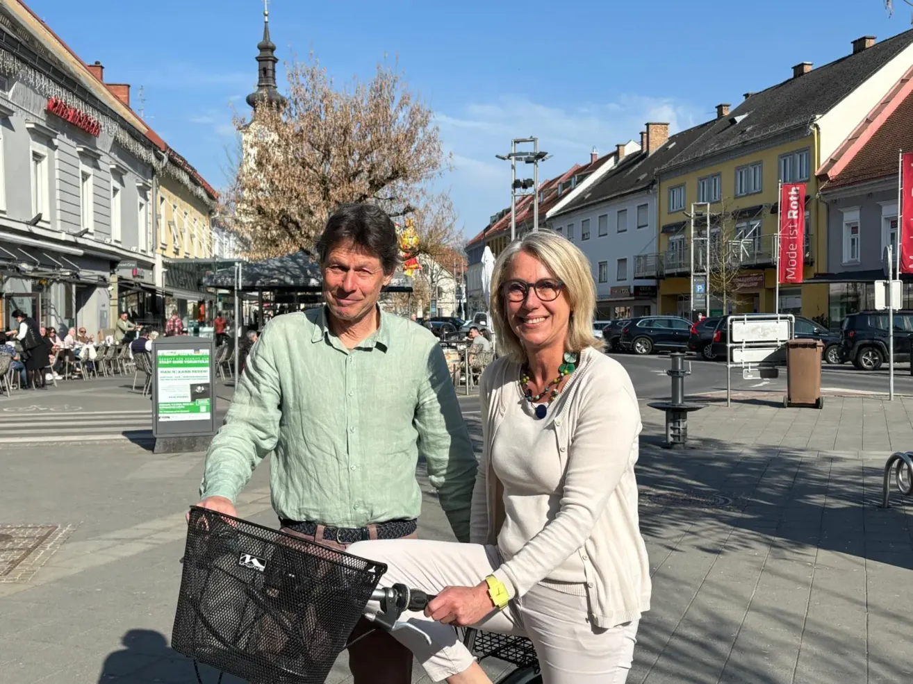 Ein Mann und eine Frau posieren für ein Foto in einer urbanen Umgebung. Sie halten ein Fahrrad, und der Mann trägt ein grünes Hemd. Hinter ihnen befindet sich eine Stadtlandschaft mit Gebäuden und Bäumen.