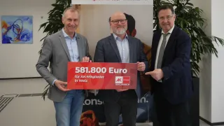 Drei Männer stehen vor einem Plakat und halten ein großes rotes Schild mit der Aufschrift 581.800 Euro für AK-Mitglieder in Weiz.