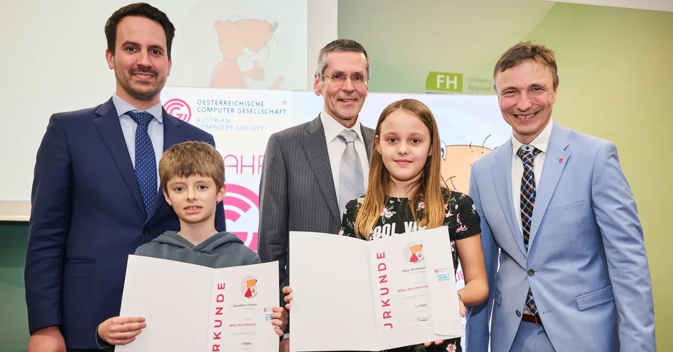 Ein Mann und eine Frau überreichen zwei Kindern, einem Jungen und einem Mädchen, die zusammenstehen, Urkunden. Der Mann trägt Anzug und Krawatte, und die Frau trägt ein Blumenhemd. Der Junge trägt eine Kapuzenjacke, und das Mädchen trägt ein schwarzes Shirt mit Blumendesign. Sie stehen vor einer Wand mit Text und Logos.