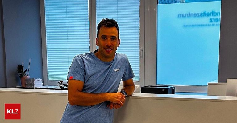 Ein Mann steht in einem Büro an einem Fenster. Er lächelt und trägt ein blaues T-Shirt. Hinter ihm ist ein Fenster mit Jalousien und ein schwarzer Drucker zu sehen.