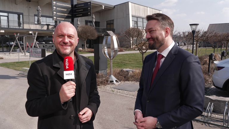 Zwei Männer stehen draußen. Einer hält ein Mikrofon und lächelt. Dahinter ist ein Rasen mit einer Metallstruktur und ein Gebäude mit Schildern.