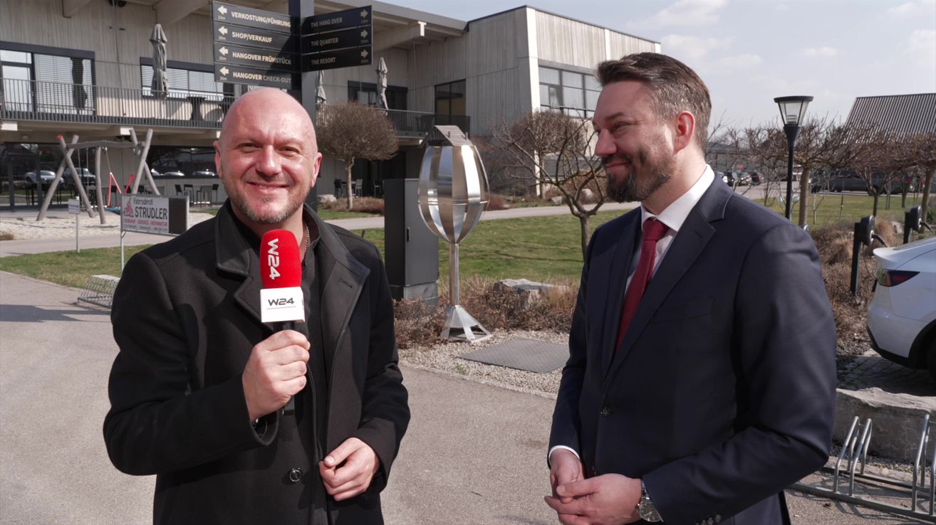 Zwei Männer stehen draußen. Einer hält ein Mikrofon und lächelt. Dahinter ist ein Rasen mit einer Metallstruktur und ein Gebäude mit Schildern.