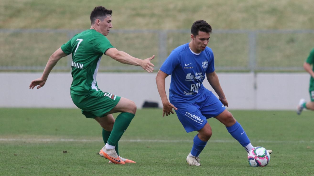 Zwei Fußballspieler in Aktion auf einem Feld, einer in Grün und der andere in Blau. Der Spieler in Grün läuft und der Spieler in Blau schaut sich den Ball an. Dahinter ist ein unscharfer Blick auf einen Zaun und ein grasbewachsenes Feld.