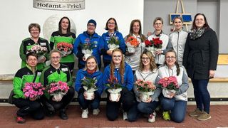 Eine Gruppe von Frauen in Sportkleidung, die mit Blumen für ein Foto posiert, mit einem Schild im Hintergrund, das 'amnt - Priel' steht.