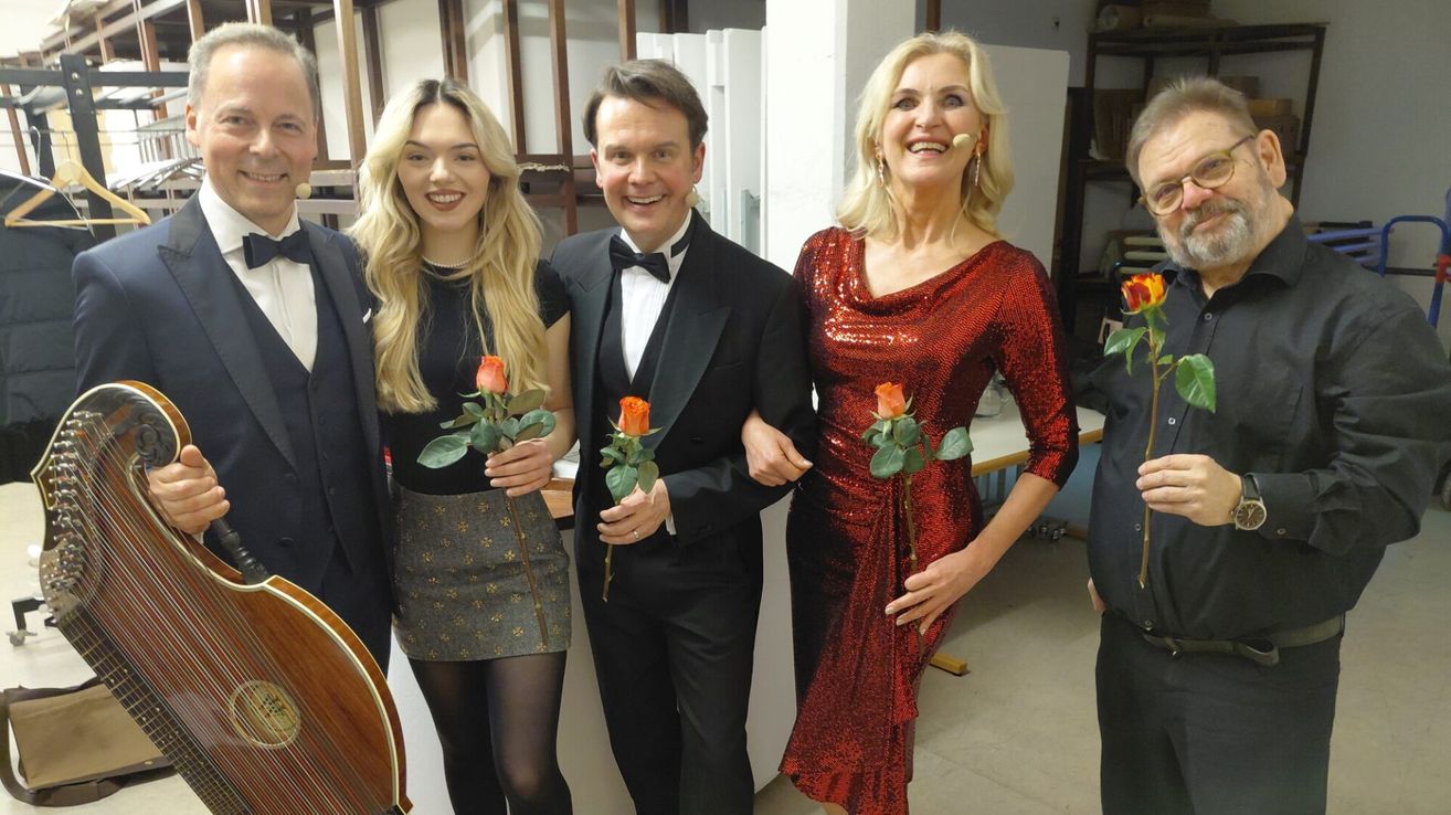 Drei Menschen in formeller Kleidung stehen zusammen und lächeln für ein Foto. Der Mann hält Rosen, und die Frauen haben Rosen in den Händen. Sie scheinen sich hinter der Bühne zu befinden.