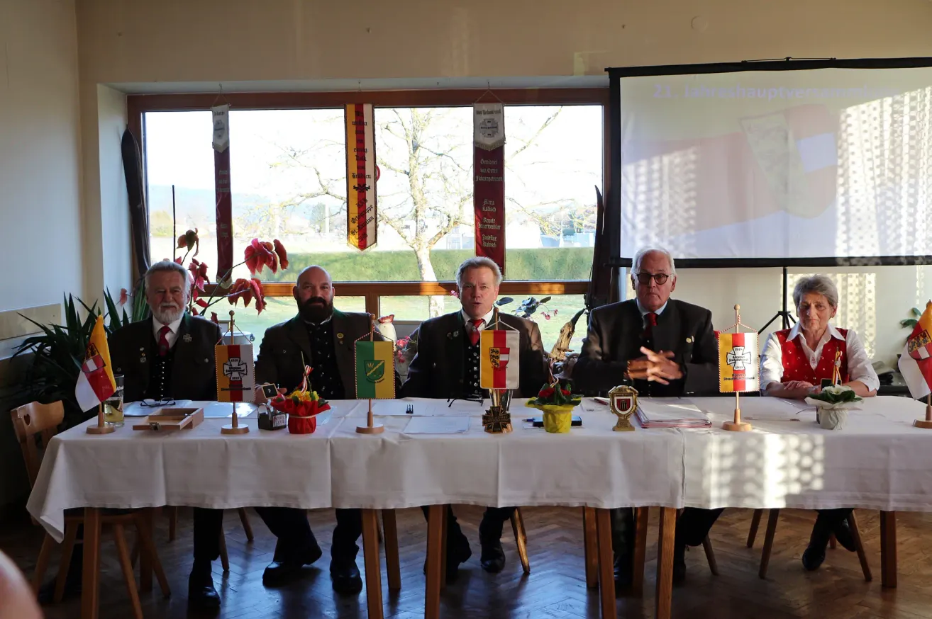 Fünf Männer in Anzügen und Krawatten sitzen an einem Tisch mit Flaggen und Blumen vor ihnen. Hinter ihnen ist eine Projektionsleinwand sichtbar.