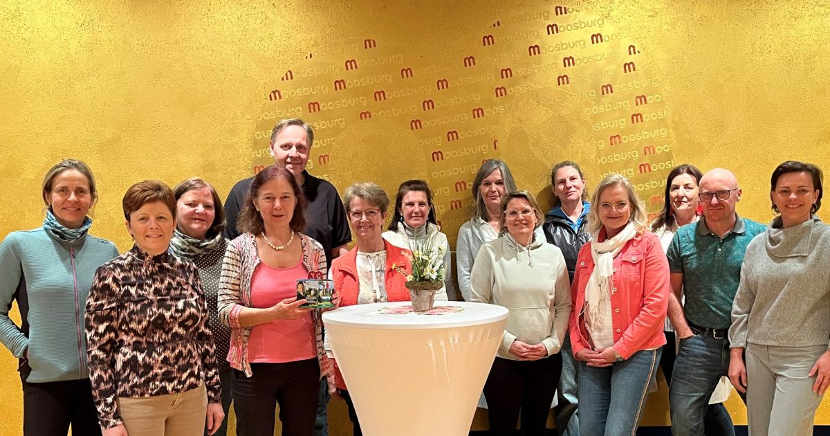 Eine Gruppe von Frauen und ein Mann posiert für ein Foto vor einer gelben Wand. Eine Frau hält eine kleine Schachtel und Blumen.