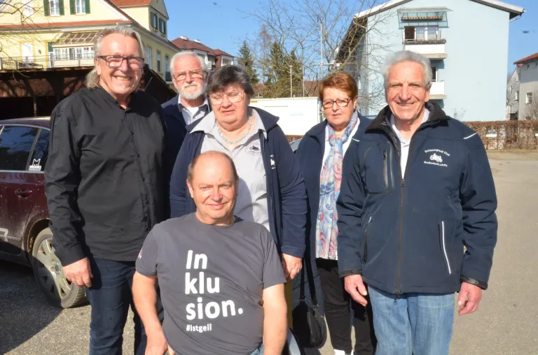 Eine Gruppe von Menschen steht vor einem Gebäude. Ein Mann trägt ein graues T-Shirt mit den Worten 'In klu sion' und '#1stgeil'. Der Mann auf der rechten Seite hat eine blaue Jacke mit weißen Aufschriften.