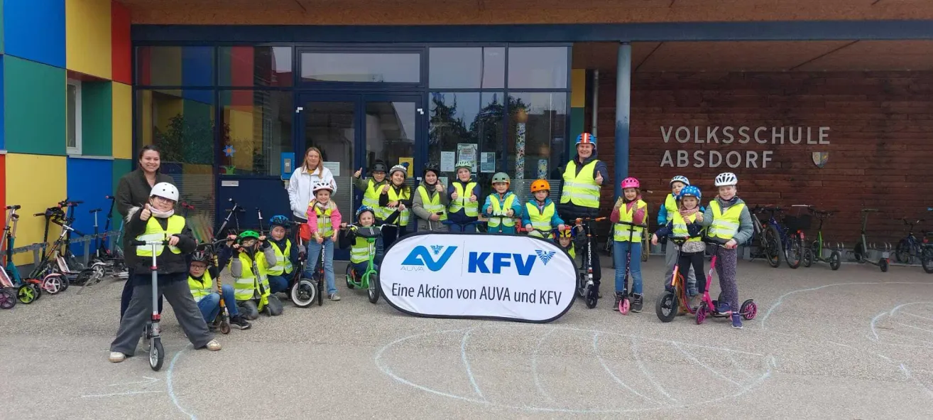 Kinder und Erwachsene in Sicherheitswesten posieren für ein Foto vor einem Gebäude mit einem Banner, das 'Eine Aktion von AUVA und KFV' besagt.