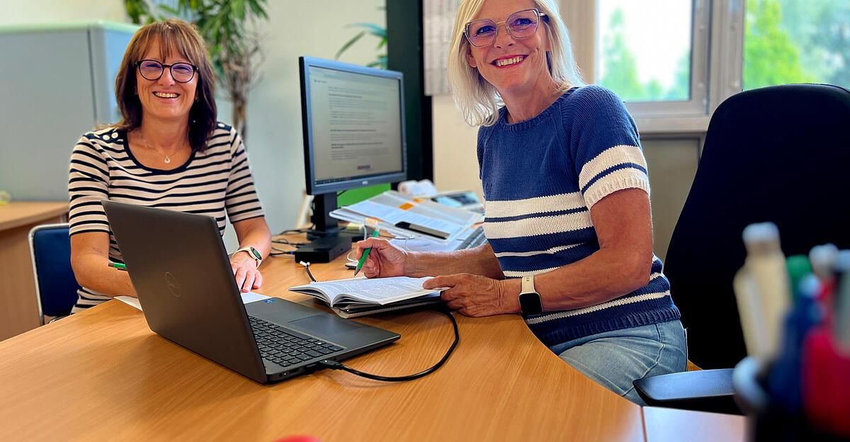 Zwei Frauen sitzen an einem Schreibtisch, eine arbeitet an einem Laptop und die andere lächelt. Sie sind von Bürogeräten umgeben.