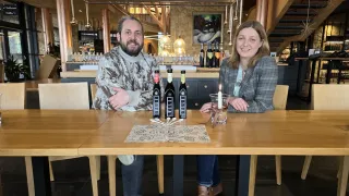 Ein Mann und eine Frau sitzen an einem Tisch mit drei Flaschen Öl und einer Kerze. Sie sind in einem Restaurant mit Holztischen und Stühlen. Hinter ihnen steht eine Theke mit Weinflaschen und einer Tafel mit Schrift.