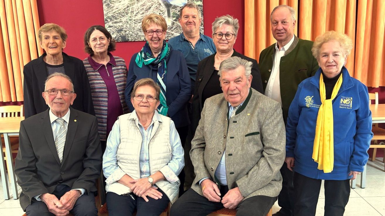 Eine Gruppe von Personen, einschließlich Männer und Frauen, posiert für ein Foto in einem Raum. Sie tragen alle eine Brille und lächeln. Der Mann in der Mitte sitzt auf einem Hocker und scheint in die Kamera zu schauen.