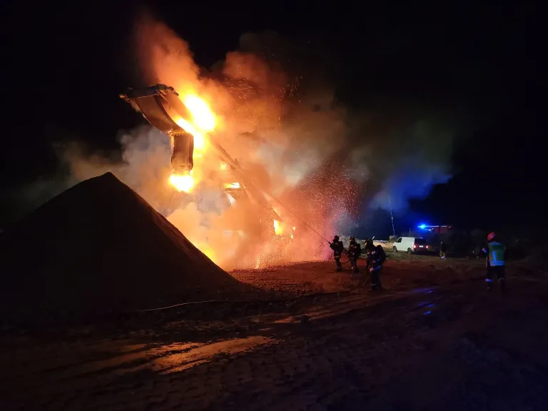 Firefighters extinguish a burning vehicle at night, with smoke and flames rising from it. Nearby, a pile of dirt and a white van are visible.