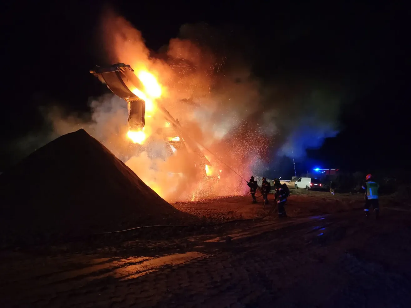 Firefighters extinguish a burning vehicle at night, with smoke and flames rising from it. Nearby, a pile of dirt and a white van are visible.