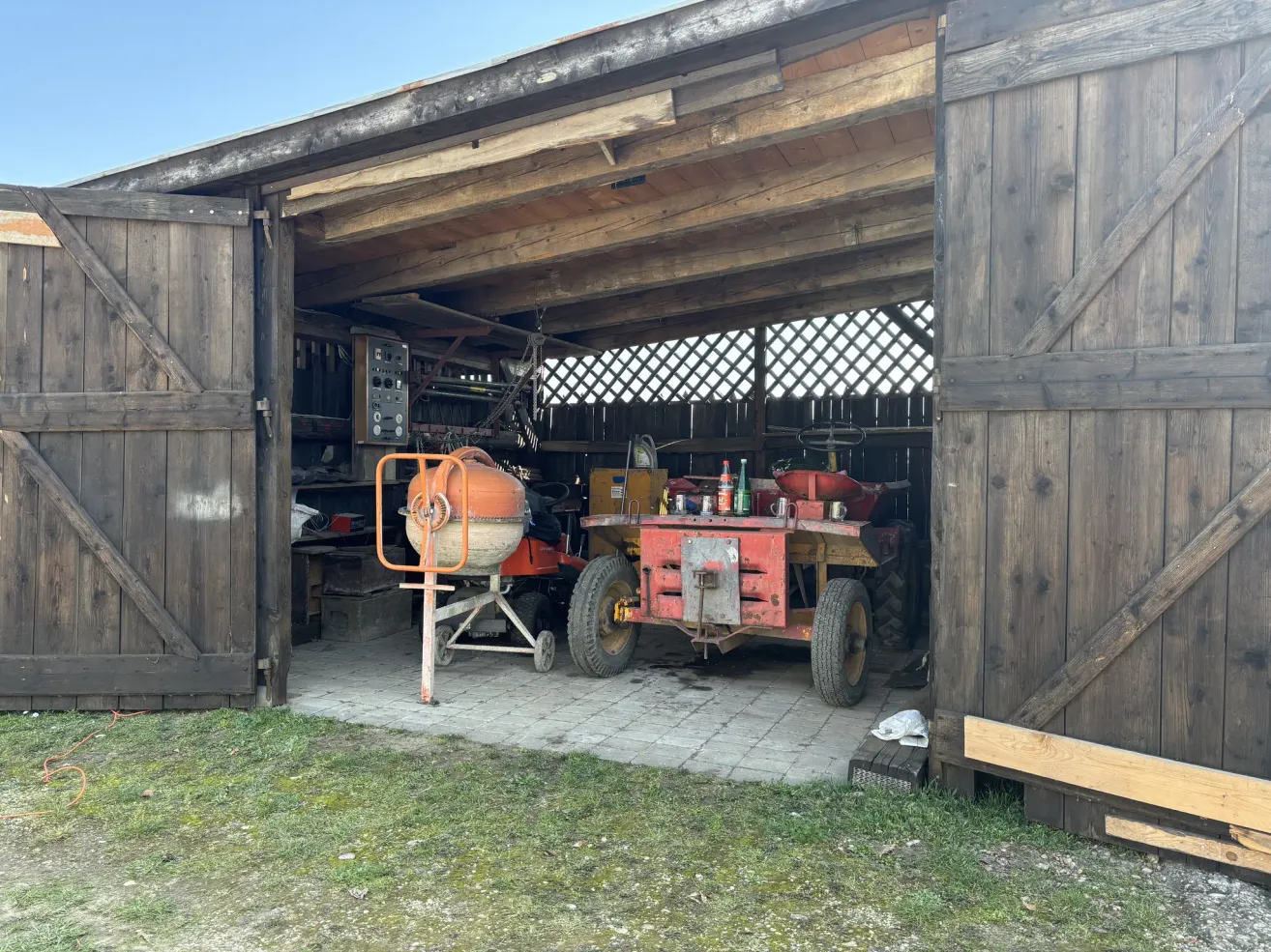 Eine Scheune mit zwei Traktoren, von denen einer mit einem Zementmischer ausgestattet ist, steht offen mit einer Holztür und zeigt eine Mischung aus landwirtschaftlichen Werkzeugen und Ausrüstung.