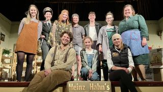 Eine Gruppe von Menschen in Kostümen steht und sitzt auf einer Bühne. Ein Schild unten lautet 'Heinz Hahn's'.