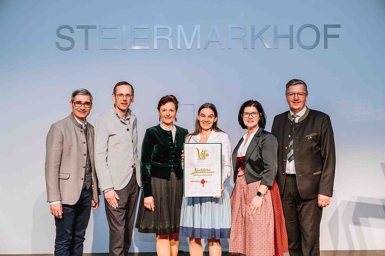 Fünf Personen stehen gemeinsam vor einem Banner mit der Aufschrift 'Steiermarkhof'. Sie lächeln und scheinen für ein Foto zu posieren. Sie halten ein Zertifikat mit den Worten 'Kandidatinnen' und 'Vollwertige und Markus'.