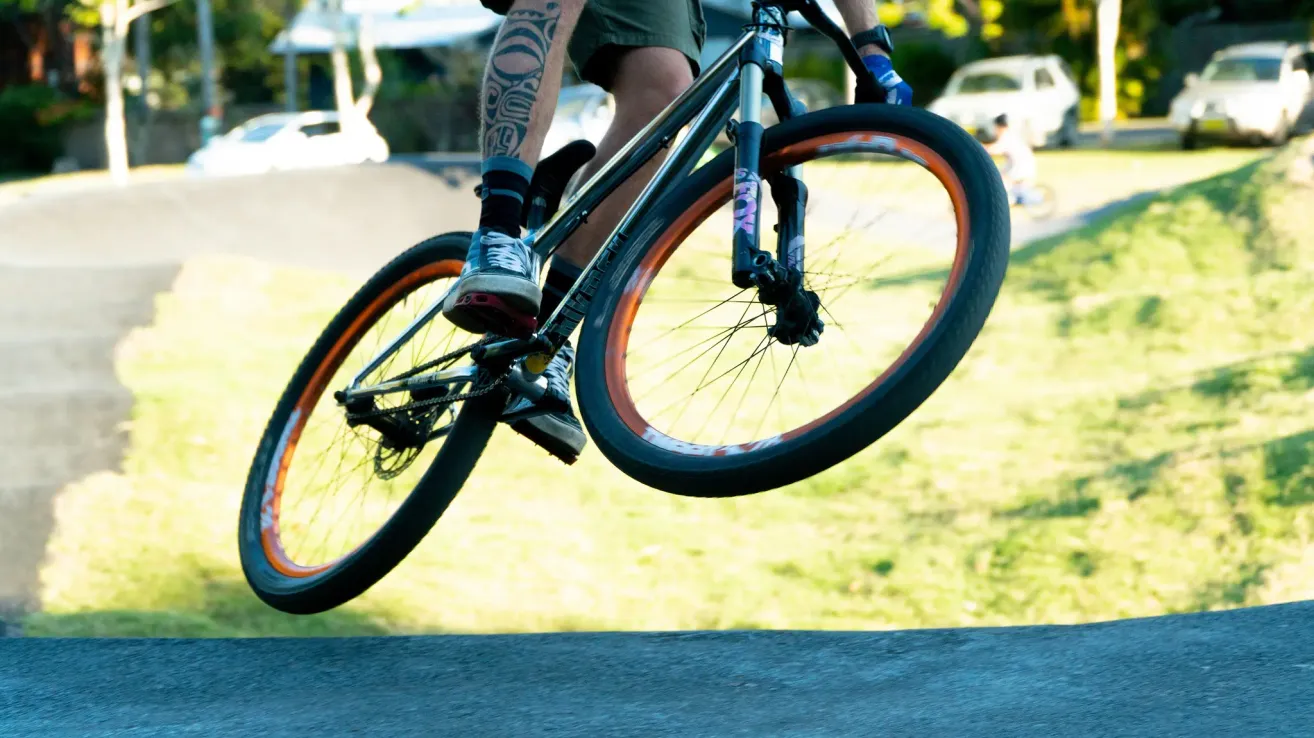 Eine Person auf einem Fahrrad mit orangefarbenen Rädern führt einen Stunt auf einer Rasenfläche aus. Der Fahrer hat ein Tattoo am Bein.