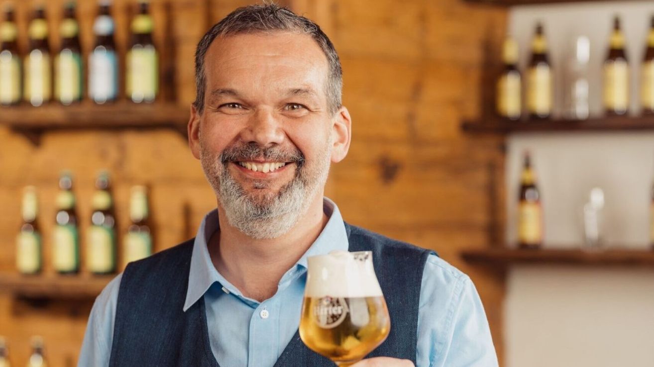 Ein lächelnder Mann mit Bart hält ein Glas Bier in der Hand und steht vor einem hölzernen Hintergrund.