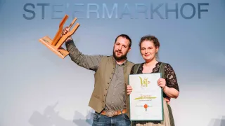 Ein Mann und eine Frau stehen auf einer Bühne, halten eine Trophäe und ein Zertifikat. Der Mann trägt eine Weste und Jeans, und die Frau trägt ein Kleid. Sie lächeln und posieren für ein Foto. Hinter ihnen ist eine Wand mit dem Text 'TEIERMARKHO'.