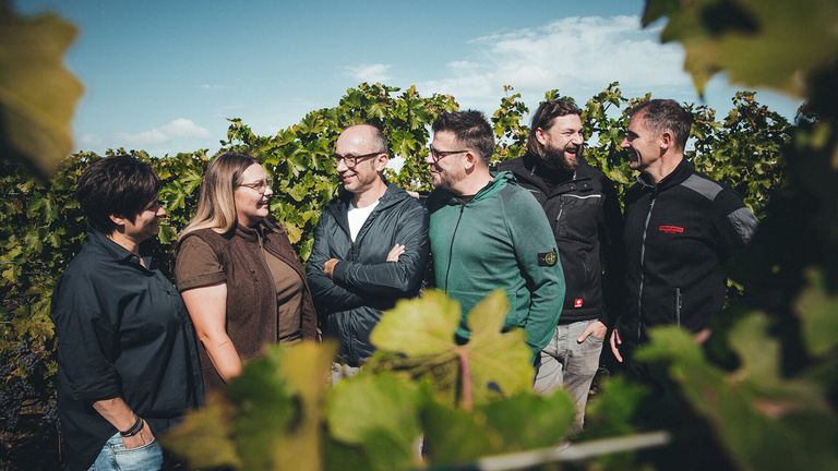 Eine Gruppe von fünf Personen steht in einem Weinberg, lächelt und posiert für ein Foto. Der Weinberg ist von grünem Laub umgeben.