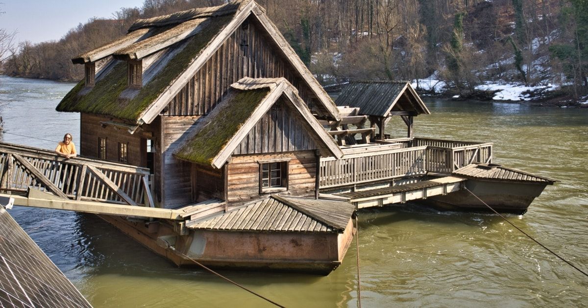 Ein altes Holzhaus ist an einem Fluss mit einer Holzbrücke und einem Deck verankert, umgeben von kahlen Bäumen und schneebedecktem Boden.