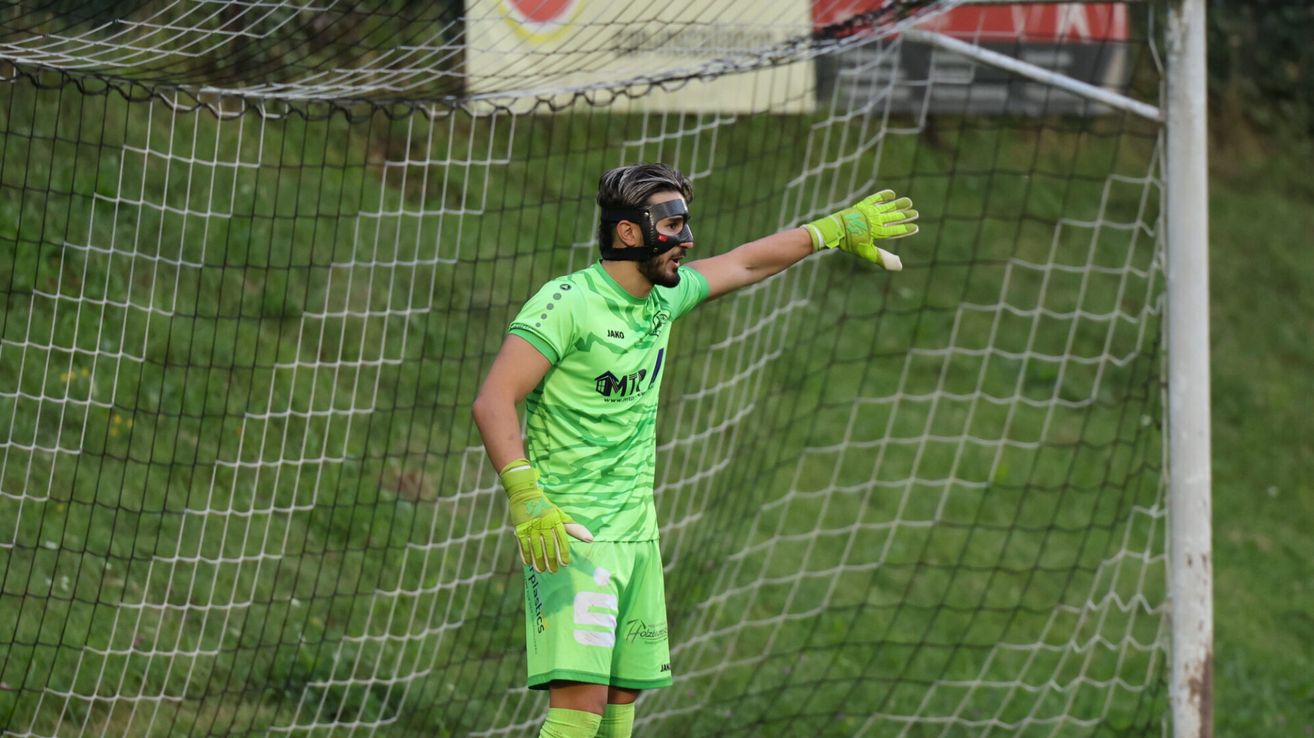 A soccer goalie in green uniform with black face mask stands near a net on a grassy field.