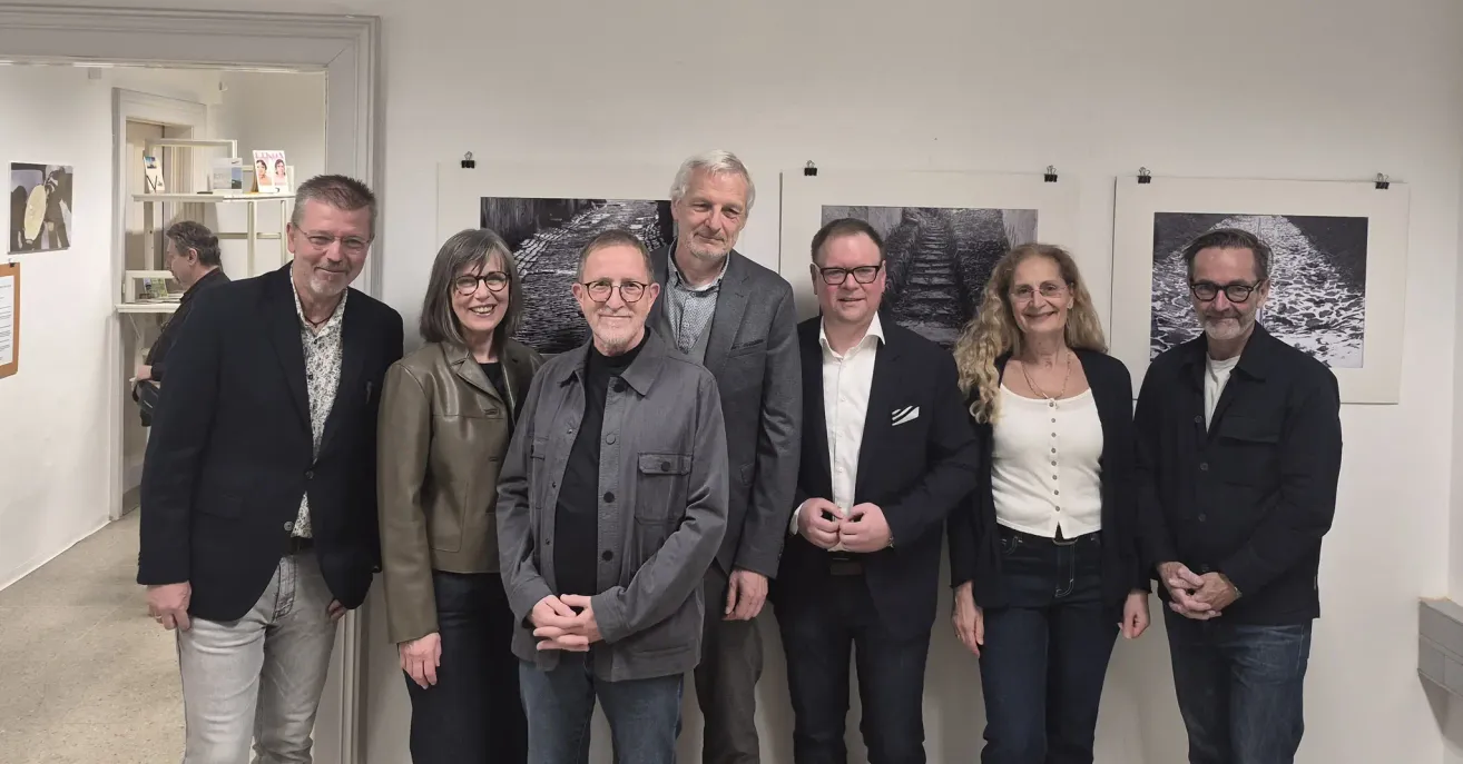 Eine Gruppe von fünf Personen steht vor einer weißen Wand mit zwei Schwarz-Weiß-Fotos. Sie alle lächeln und scheinen für ein Foto zu posieren.