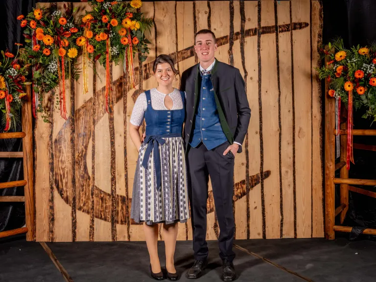 A smiling couple stands on a stage with a wooden wall in the background, both dressed in traditional attire. The woman wears a blue dress with a white apron, and the man wears a dark blue suit.