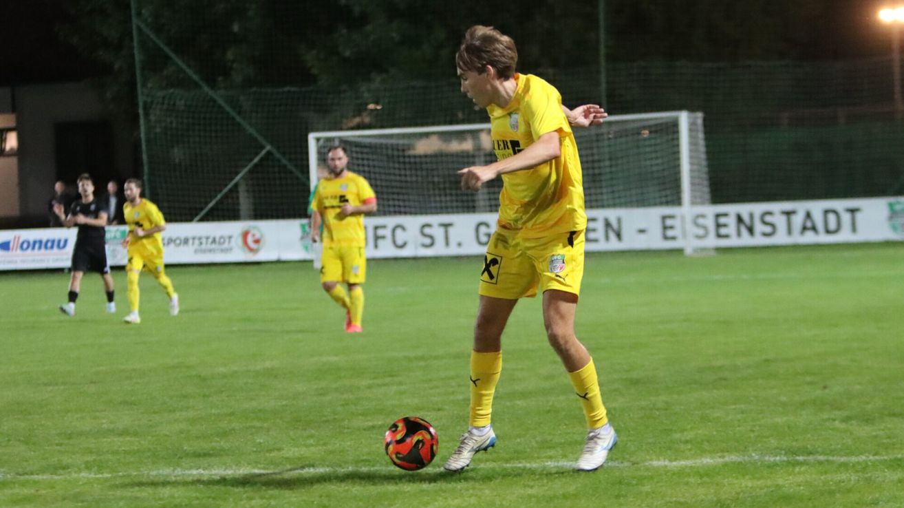 Ein Fußballspieler in einem gelben Trikot ist dabei, einen rot-schwarzen Ball zu treten. Ein weiterer Spieler in Gelb steht hinter ihm. Das Spielfeld ist von einem Zaun und einem Banner umgeben.